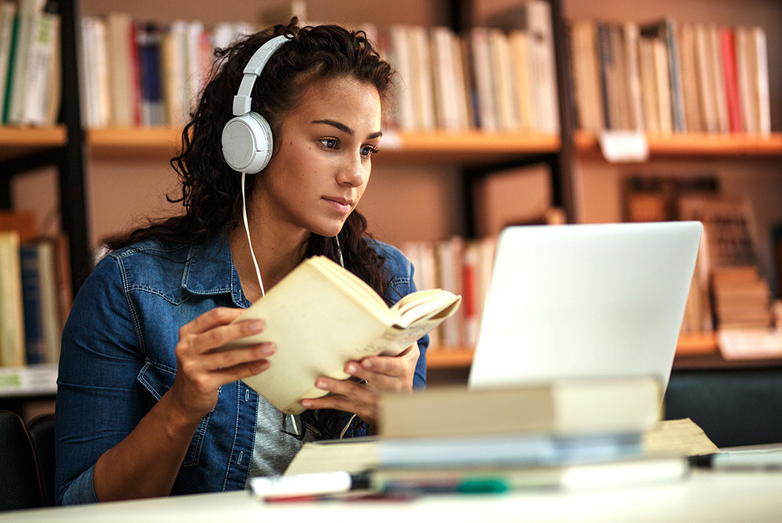 Student sitter i ett bibliotek med en bok och en bärbar dator.